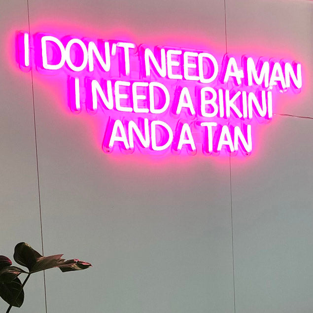 Pink neon sign featuring the words I Don't Need a Man I Need a Bikini and a Tan shown turned on against a white wall - from Custom Neon®