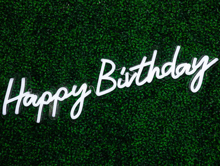 Happy Birthday Cursive Neon Sign against a green wall -photo from CustomNeon.com