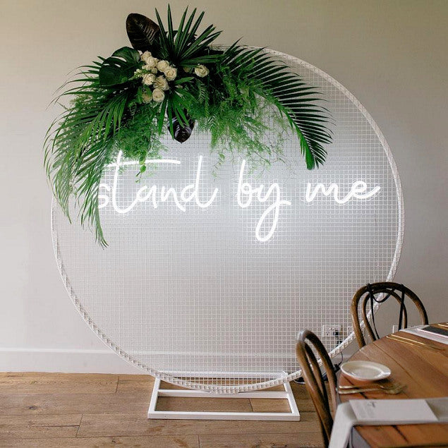 Stand by me LED neon wedding sign shown mounted on a mesh screen with a floral arrangement - photo from CustomNeon.com