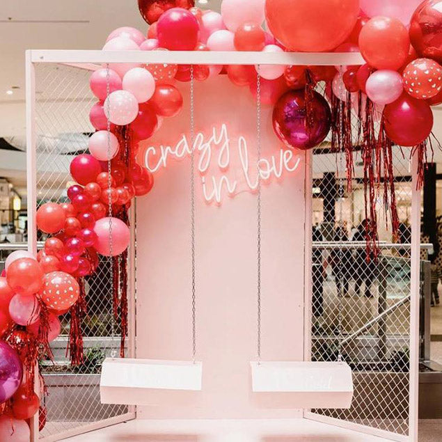 Crazy in Love neon sign wall mounted on a department store display with balloons and lover's swings - photo from Custom Neon