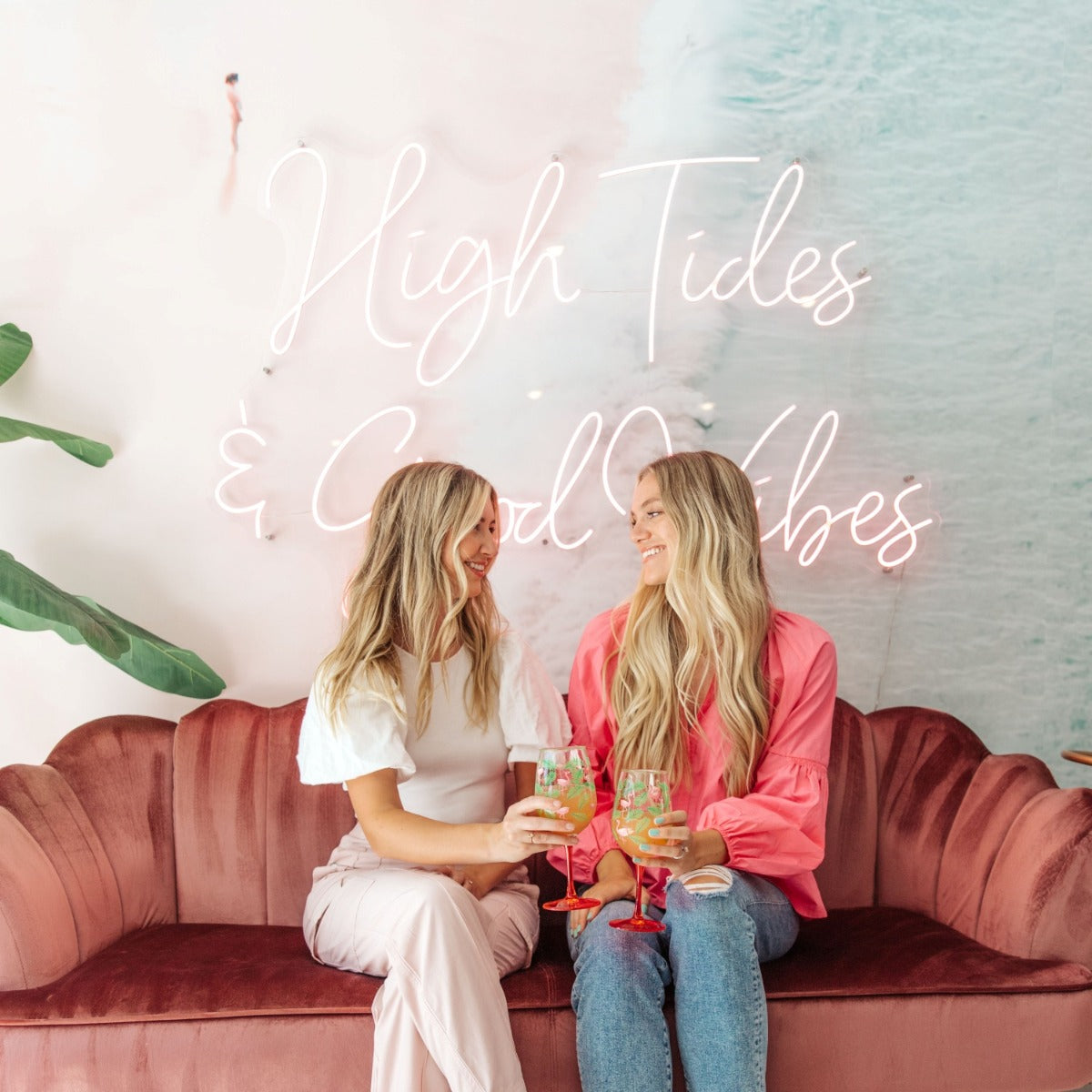 High Tides & Good Vibes pink LED neon sign installed a soft pink and blue wall mural of waves on the beach with two women holding glasses in the foreground - CUSTOM NEON™ x SOMERLED COLLECTION