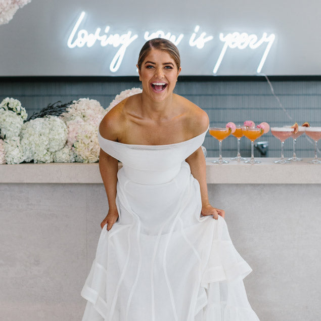 Loving you is yeezy wedding neon sign shown in white behind the bar with a beautiful bride in front - photo from CustomNeon.com